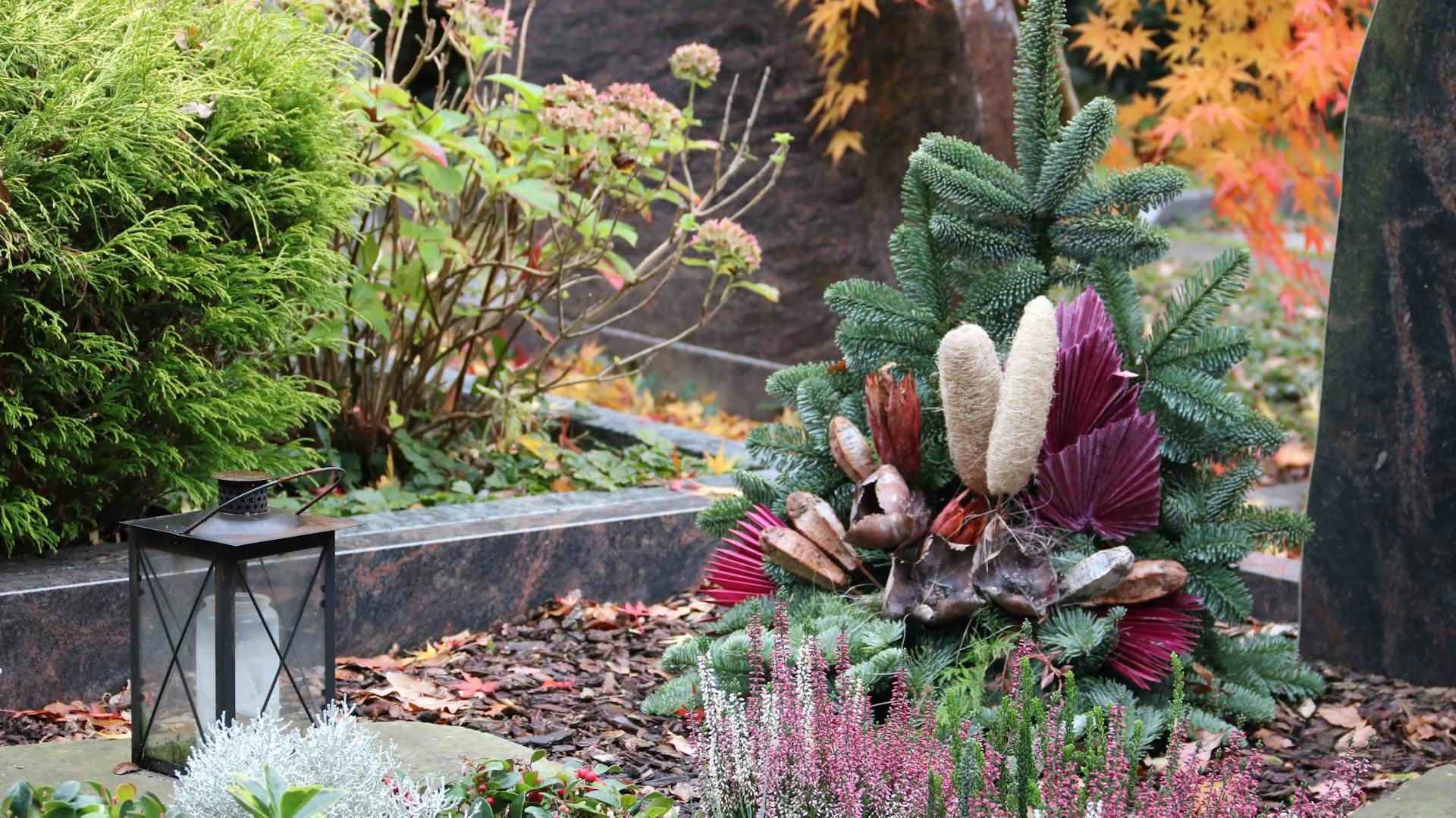Ein herbstlich bepflanztes Grab mit Rindenmulch und Grabkerze in einer schwarzen Laterne.