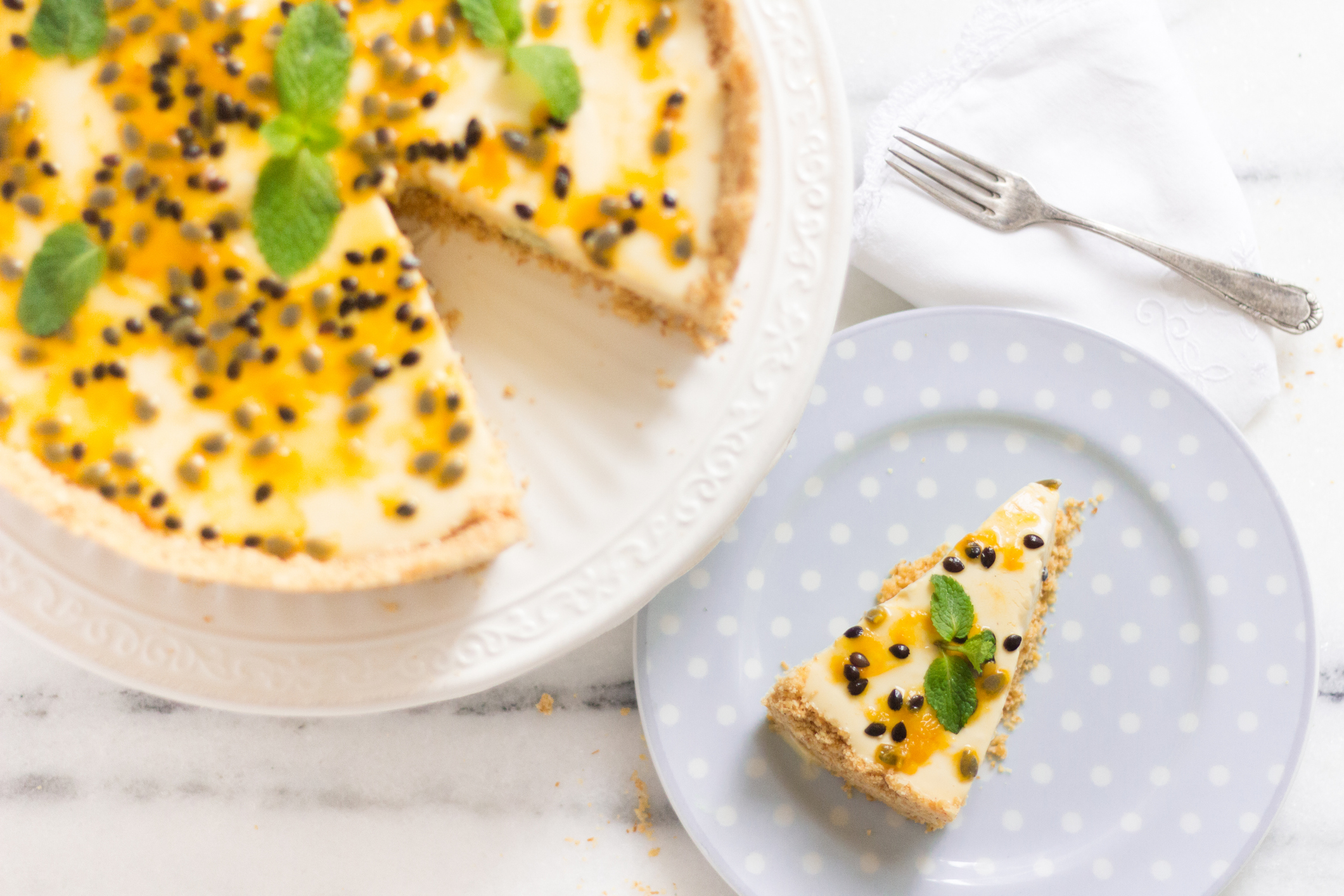 Angeschnittener Käsekuchen mit Passionsfrucht neben Kuchenteller mit einem Stück Kuchen.