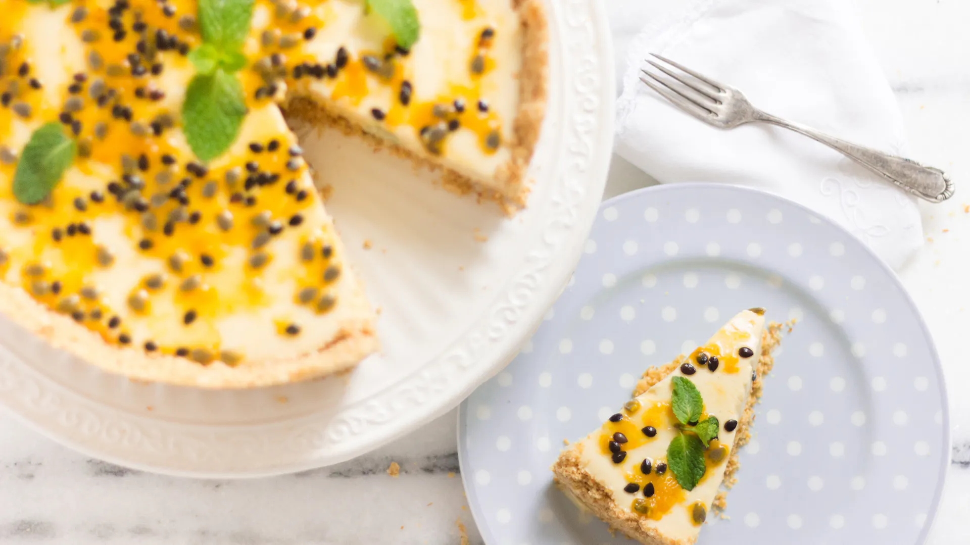 Angeschnittener Käsekuchen mit Passionsfrucht neben Kuchenteller mit einem Stück Kuchen.