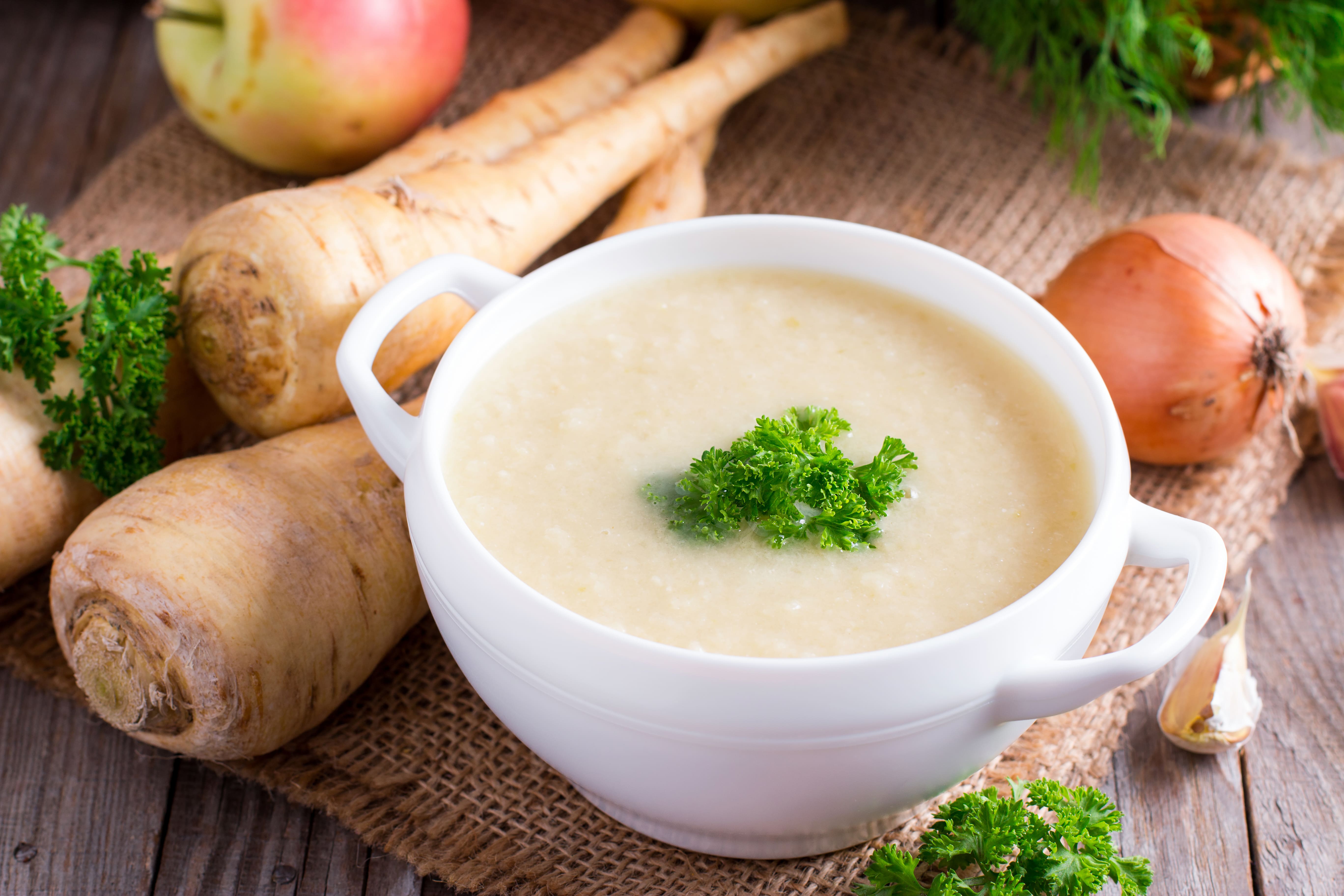 Eine Schüssel Pastinakensuppe neben Pastinaken und Zwiebeln.