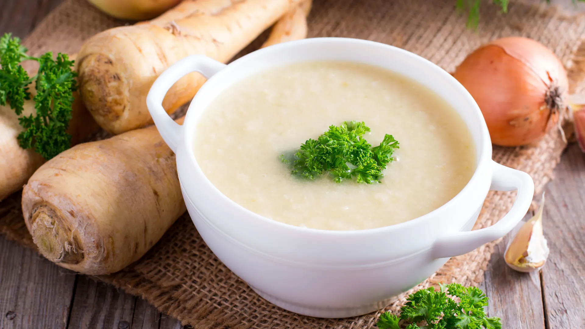 Eine Schüssel Pastinakensuppe neben Pastinaken und Zwiebeln.