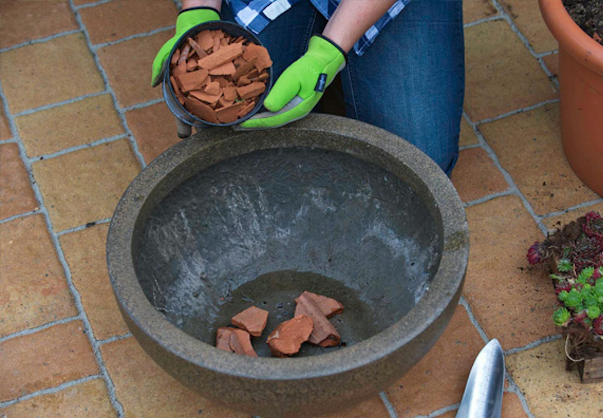 Valaki cserépdarabokat helyez el egy dézsában a vízelvezető réteg kialakításához.