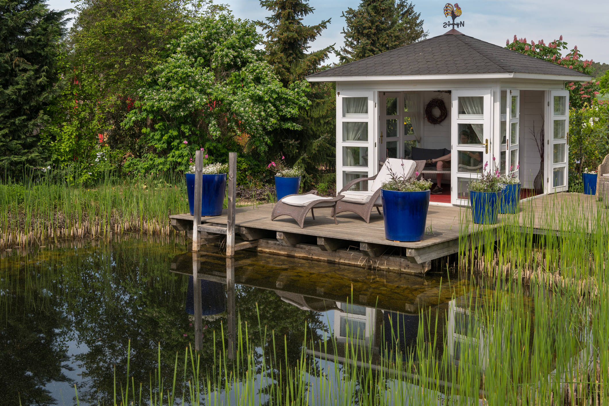 Ein mit Schilfrohr bepflanzter Gartenteich mit Gartenhaus, Steg und Gartenmöbeln.