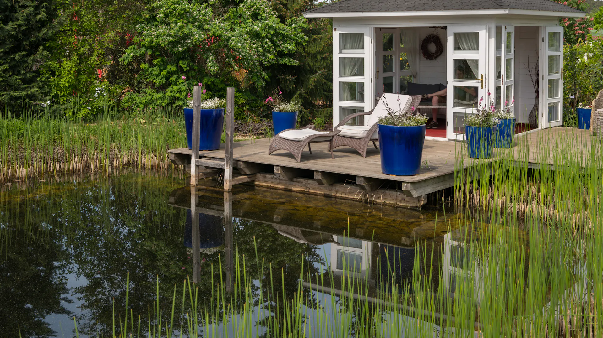 Ein mit Schilfrohr bepflanzter Gartenteich mit Gartenhaus, Steg und Gartenmöbeln.