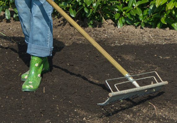 Egy gumicsizmás nő, egy bevetetlen ágyáson áll egy gereblyével.