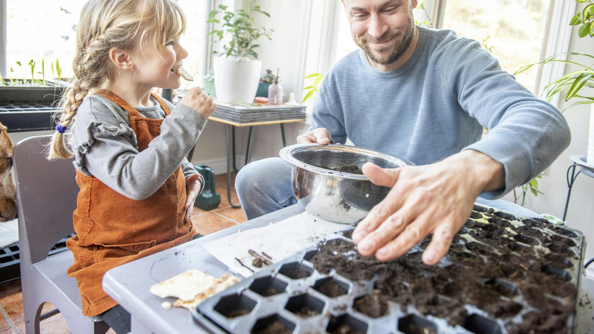 Eine Person füllt Erde in Anzuchttöpfe, ein kleines Kind sitzt daneben.
