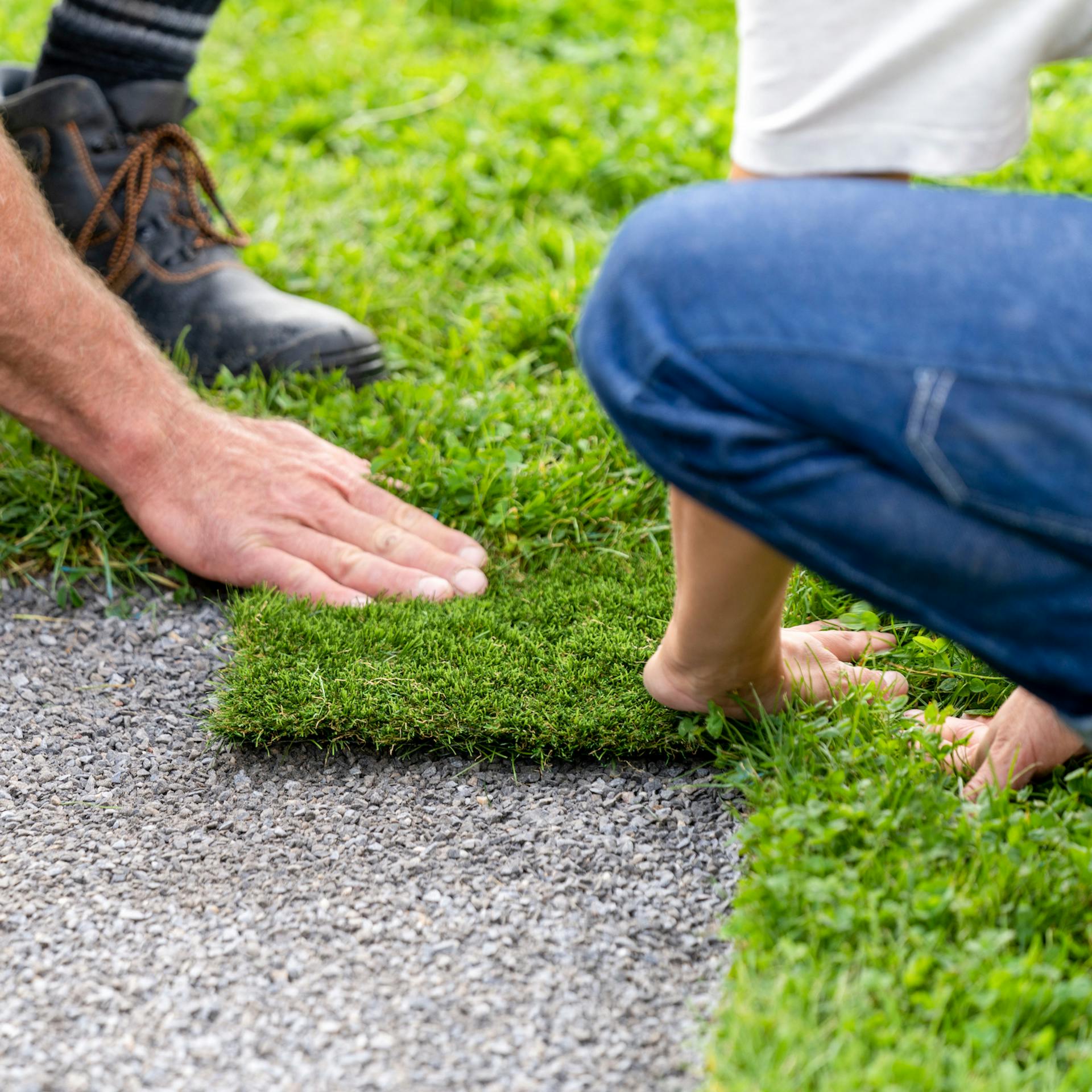 Dvě osoby drží kus umělého trávníku na písčitém podloží