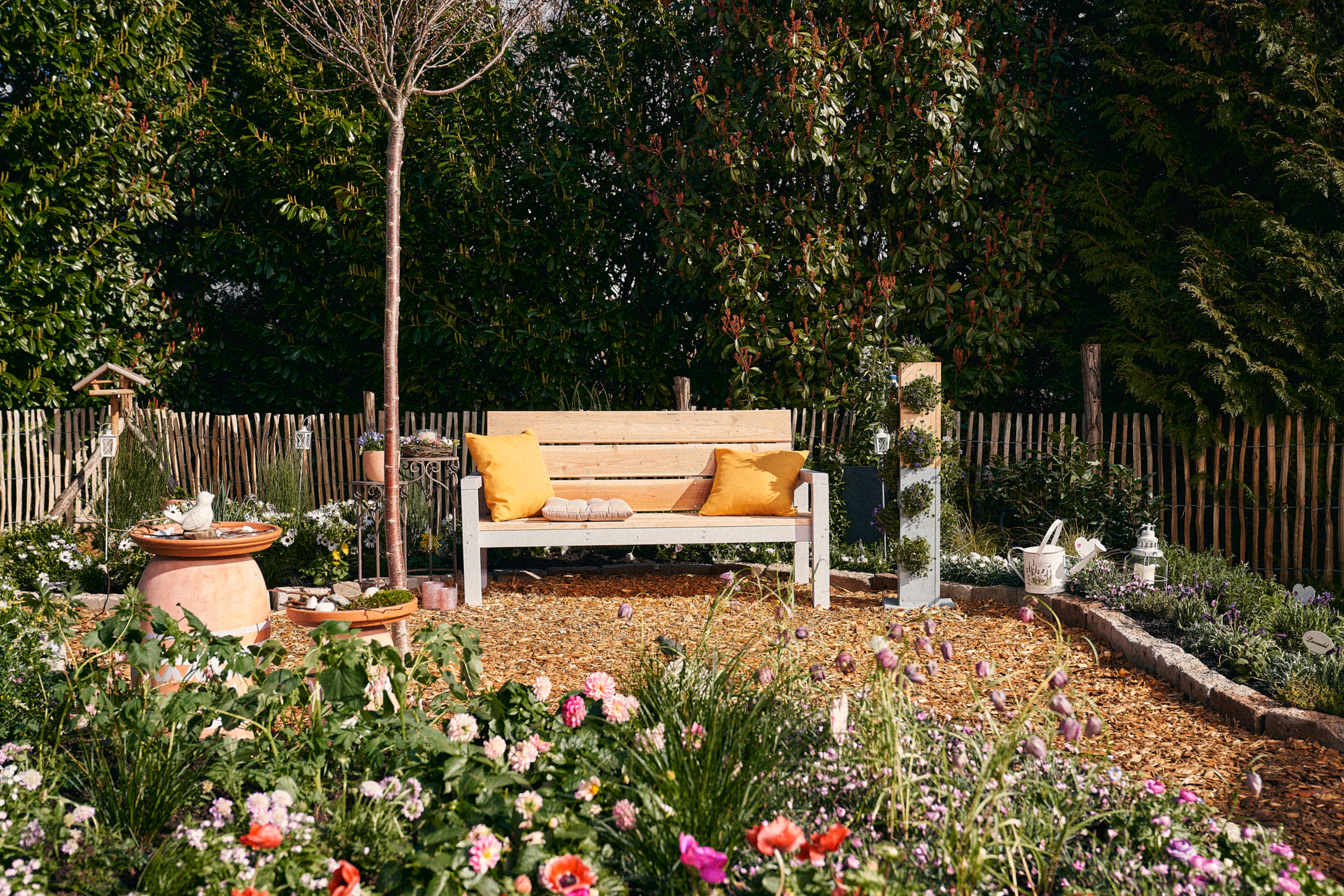 Nyáron a kertben kint áll egy pad. Mellette egy növénytorony és egy állatitató áll.