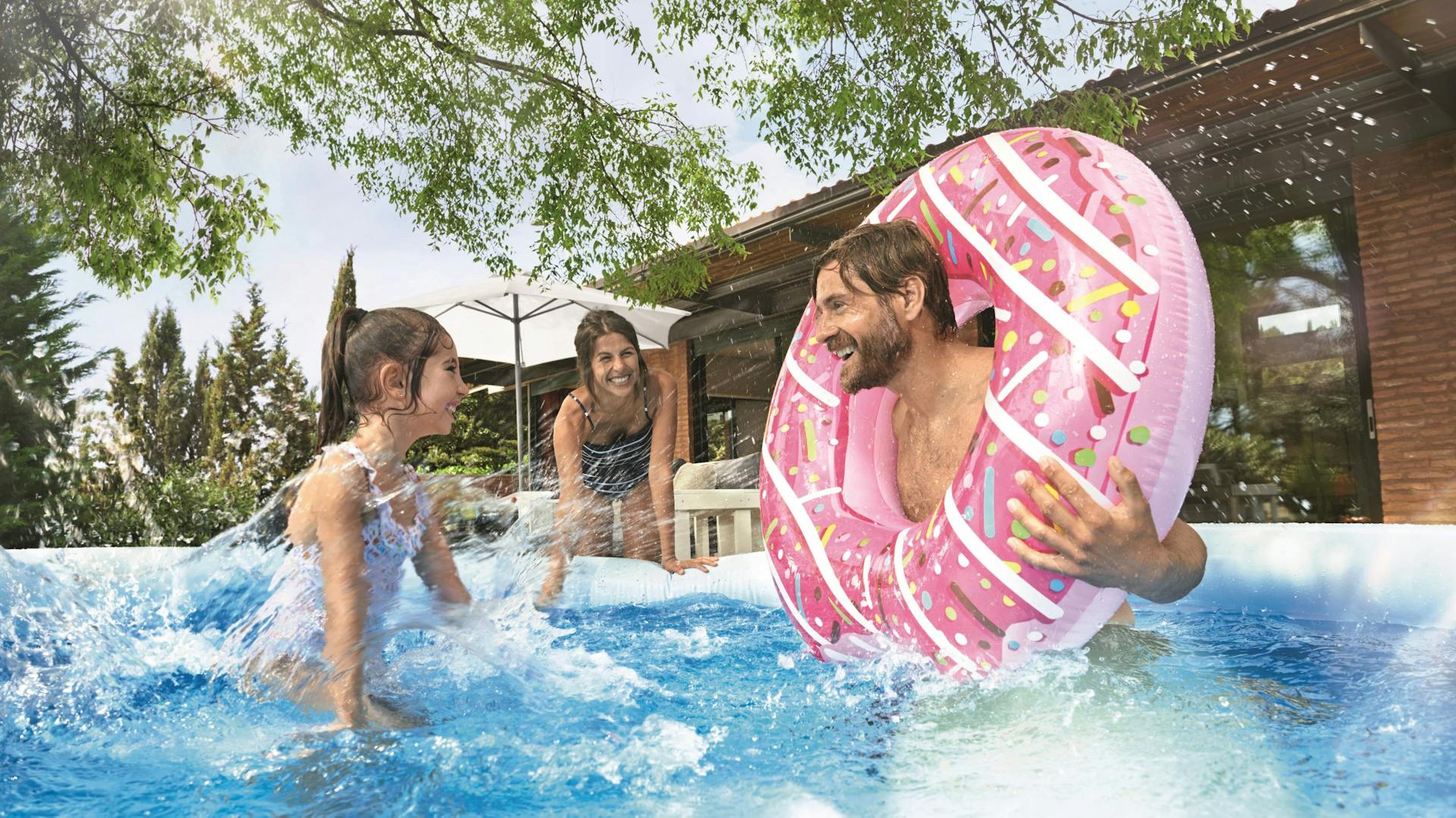 Un uomo e due bambini giocano in piscina con spruzzi d’acqua e salvagente gonfiabile.