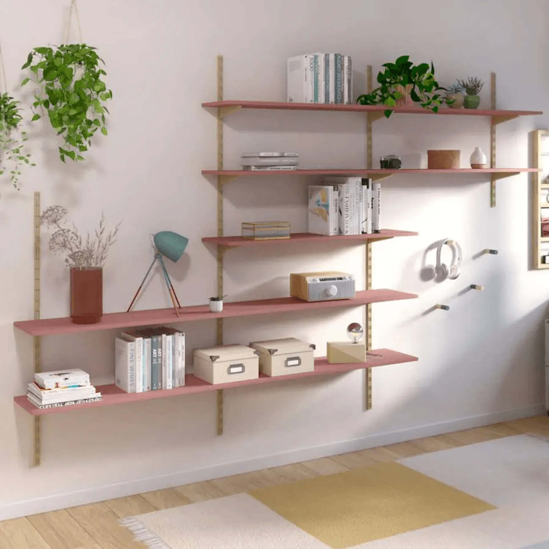 Półki ścienne z różowymi deskami zamontowane na złotych wspornikach, ozdobione roślinami, książkami, lampką i innymi dekoracjami w przytulnym, nowoczesnym pokoju.