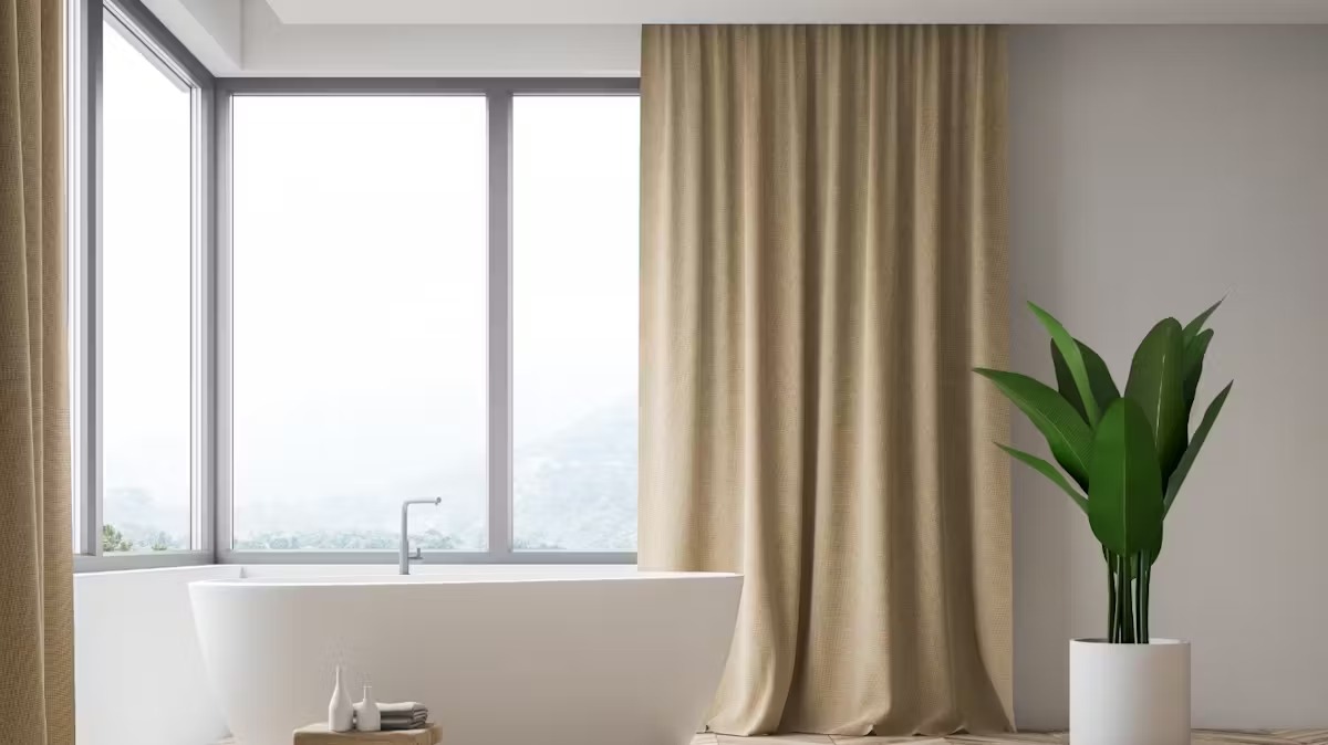 Biała wanna przy dużym, narożnym oknie, w którym wiszą beżowe zasłony.