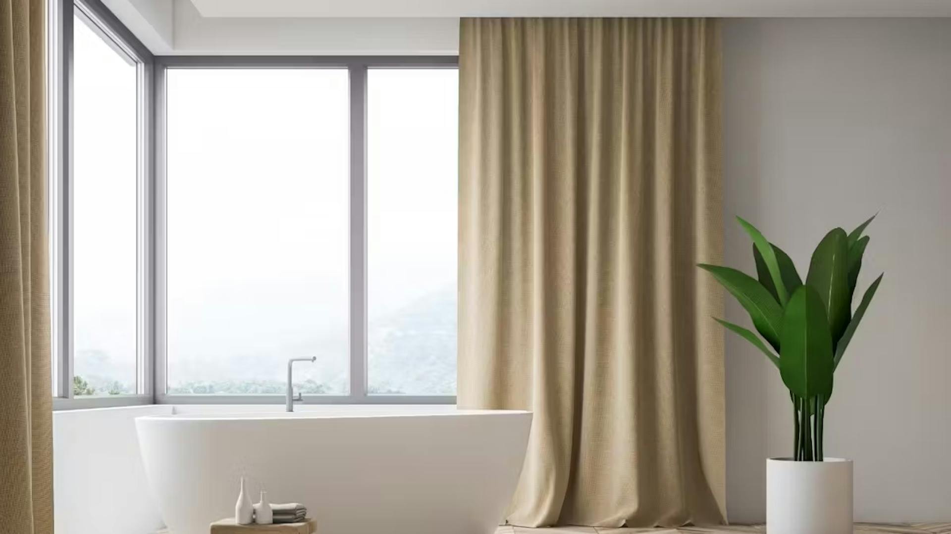 Biała wanna przy dużym, narożnym oknie, w którym wiszą beżowe zasłony.