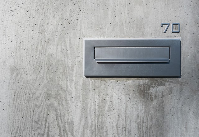 Metalowa skrzynka na listy wbudowana w betonową ścianę. Nad wlotem metalowe cyfry 7 i 0.