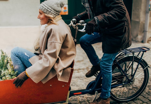 Kobieta z choinką przedniej przyczepie rowerowej. Na rowerze pedałuje mężczyzna w zimowym ubraniu.