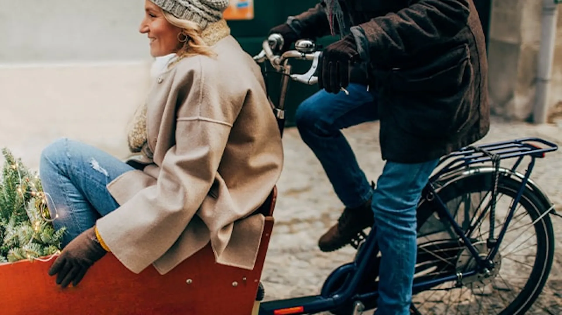 Kobieta z choinką przedniej przyczepie rowerowej. Na rowerze pedałuje mężczyzna w zimowym ubraniu.