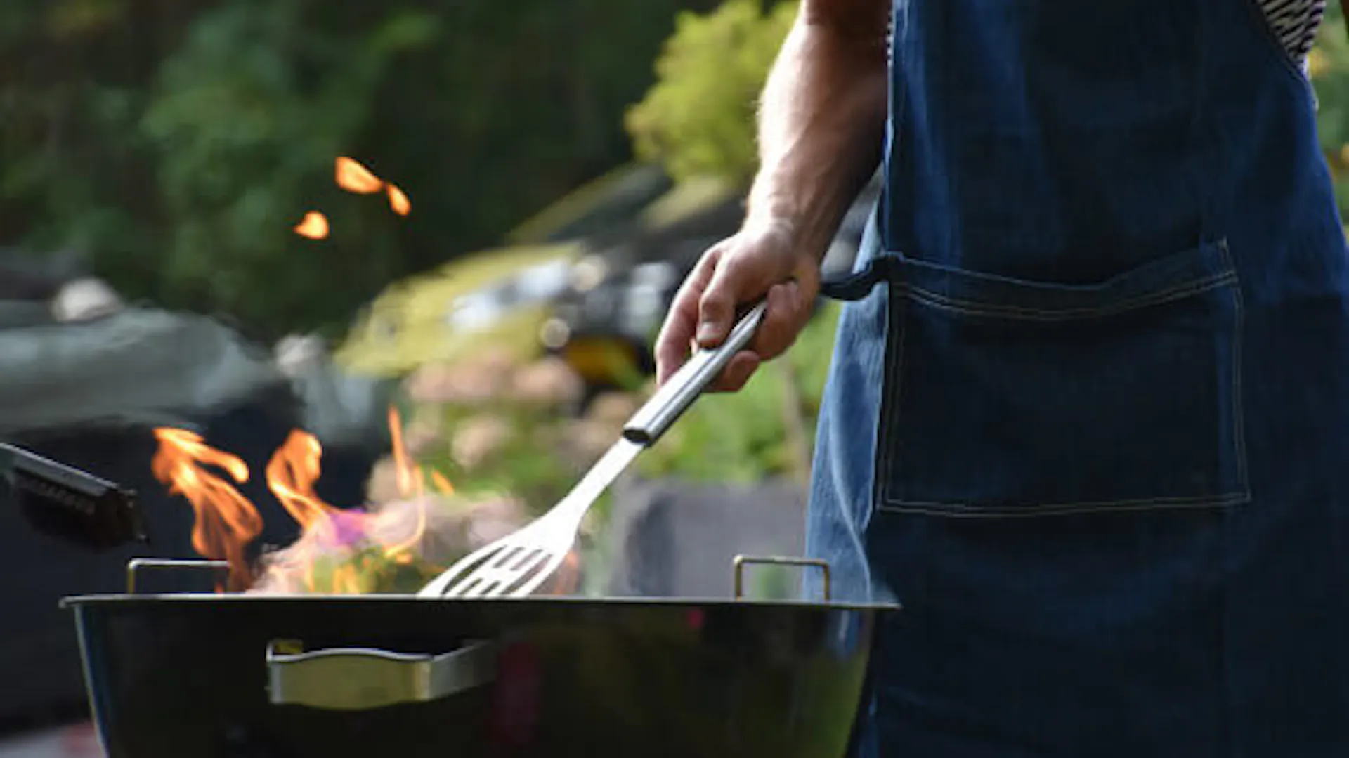 Mężczyzna w jeansowym fartuchu przy okrągłym grillu. Z grilla wydostaje się ogień.
