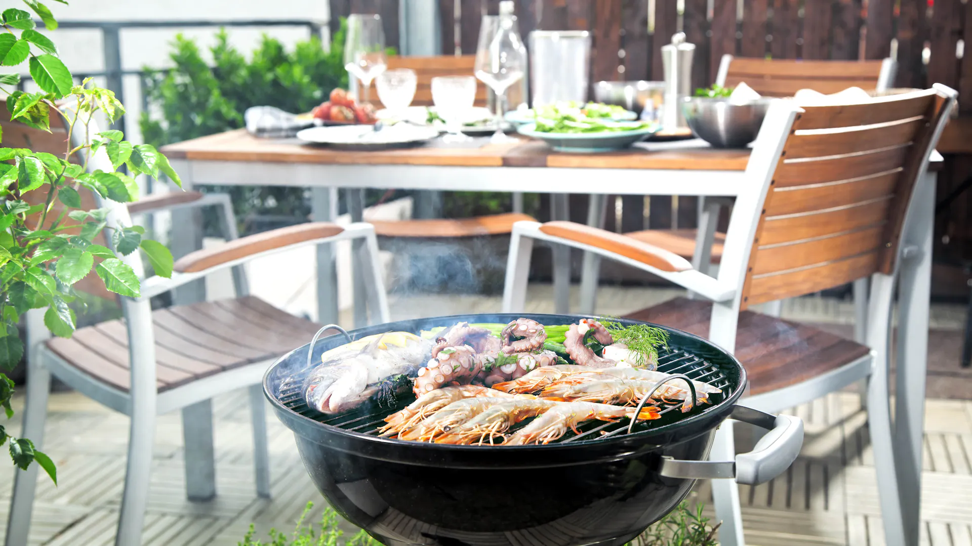 Grill na balkonie, w tle stół z zastawą, rośliny i parawan ogrodowy