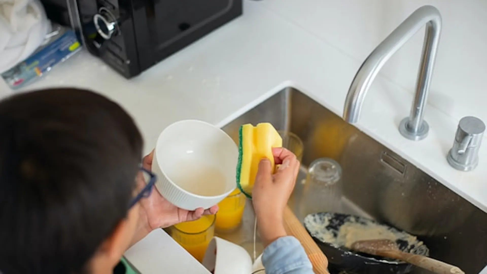 Zlew kuchenny z brudnymi naczyniami. Stojąca tyłem osoba myje gąbką białą miseczkę.