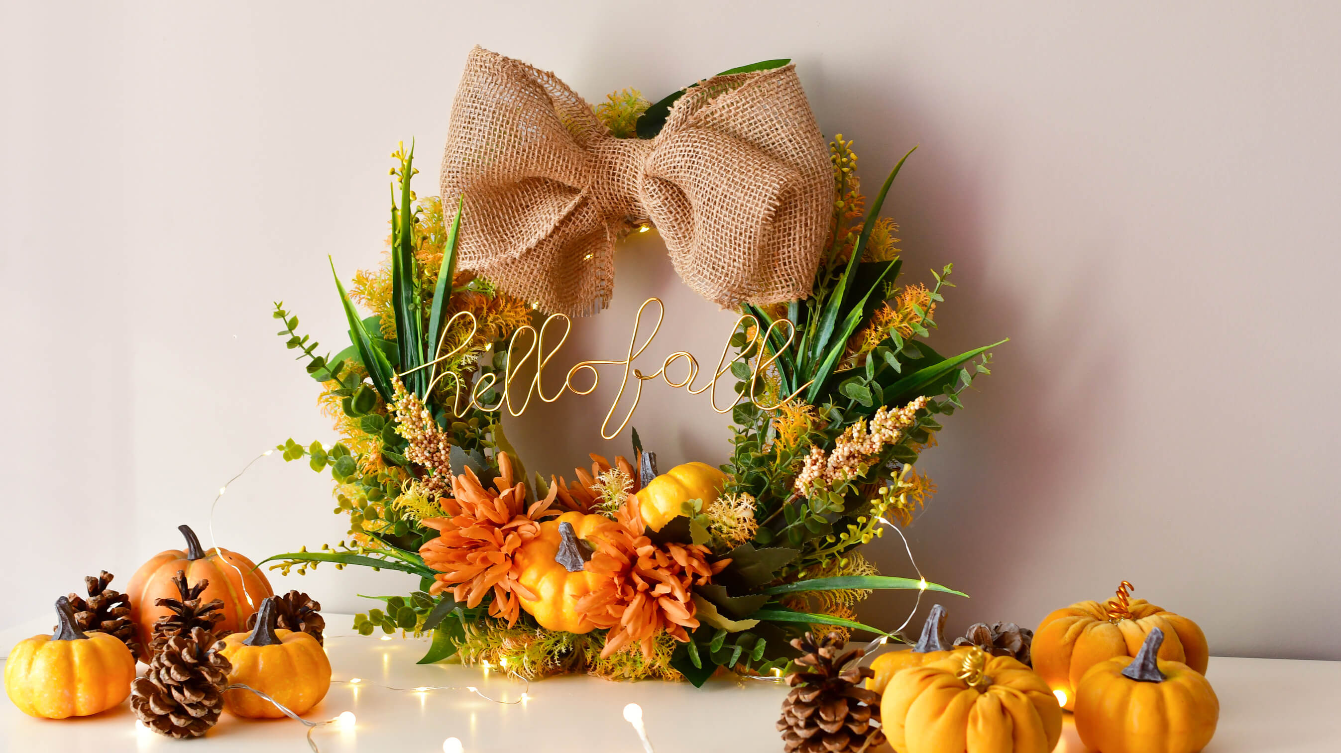 Jesienny wieniec ozdobny z napisem „hello fall”, udekorowany pomarańczowymi kwiatami, mini dyniami, zielonymi liśćmi i dużą jutową kokardą, ustawiony na białym tle z małymi dyniami i szyszkami dookoła. 