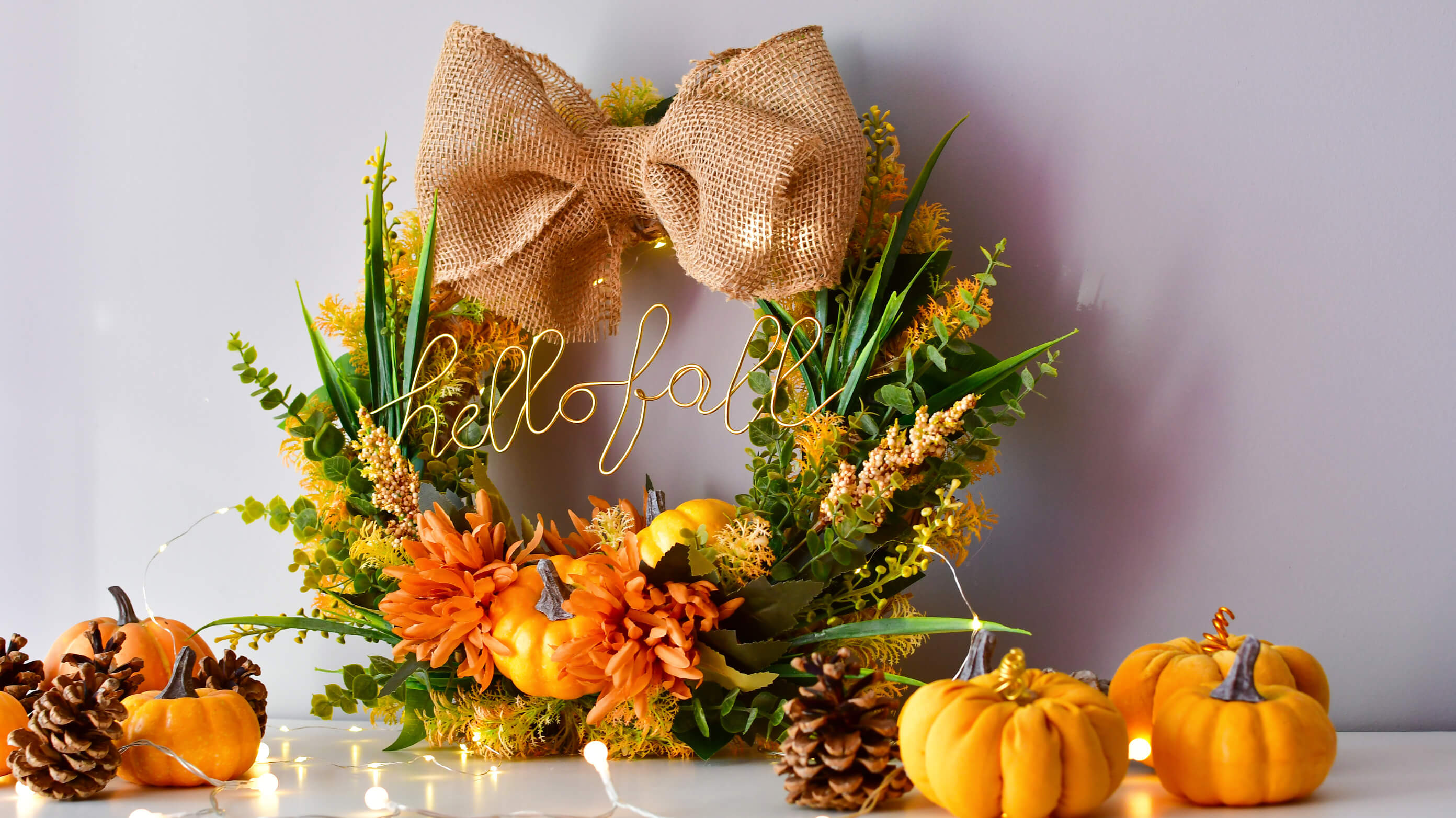 Jesienna dekoracja DIY: wianek na drzwi przygotowana samodzielnie przy użyciu mini dyni dekoracyjnych, sztucznych kwiatów, jutowej kokardy, z napisem: hello fall.