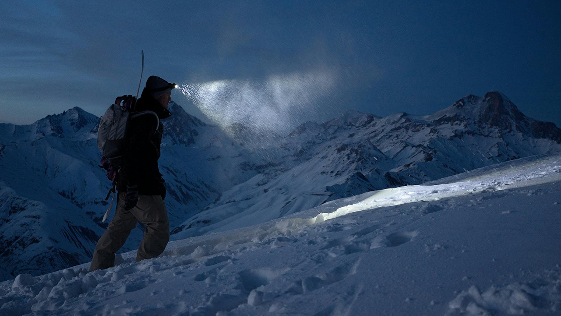 Ein Mann spaziert bei Nacht auf einem Berg durch den Schnee mit einer Ledlenser Stirnlampe