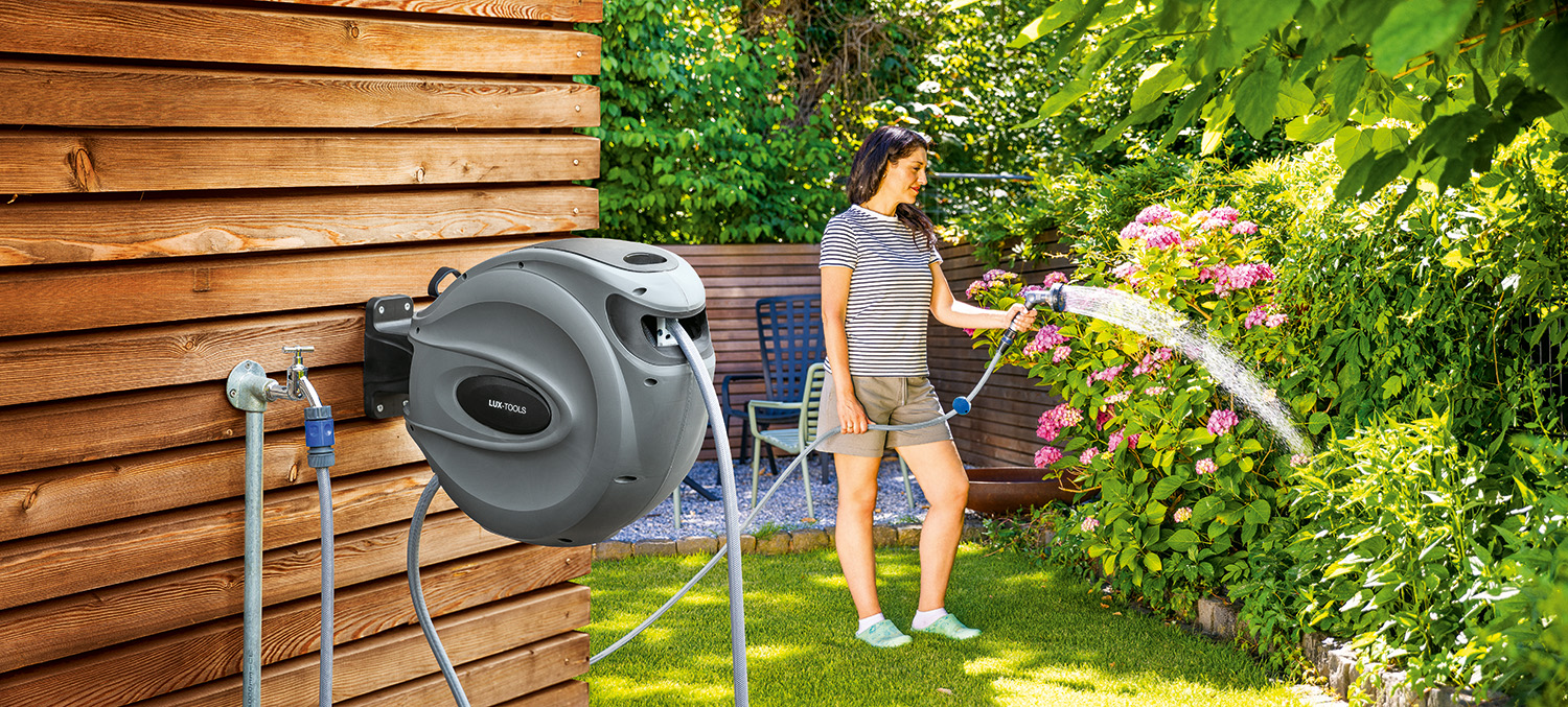 Schlauchbox die an einer Wand befestigt ist. Im Hintergund bewässert eine Frau ihren Garten