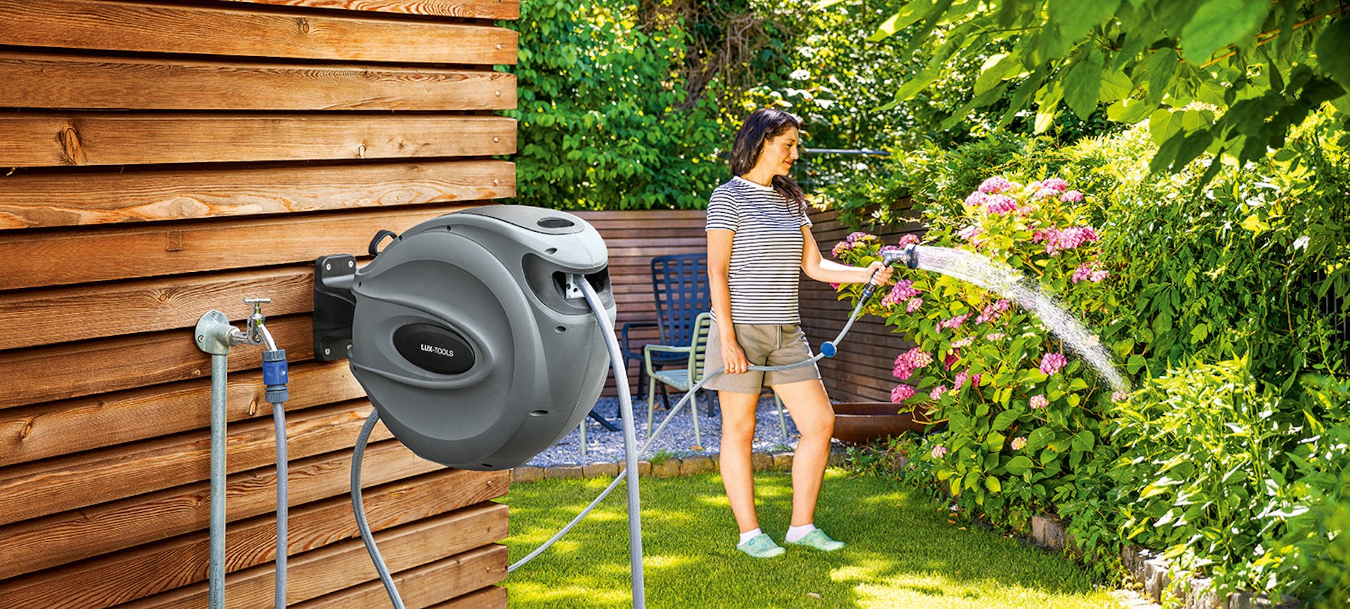 Schlauchbox die an einer Wand befestigt ist. Im Hintergund bewässert eine Frau ihren Garten